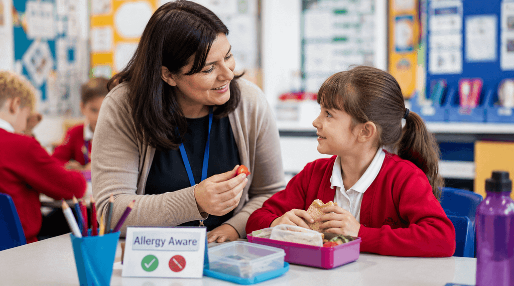 Teacher supporting a pupil during allergy awareness activities in a primary school classroom